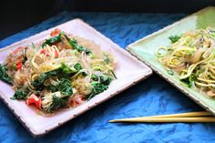 two plates with noodles, vegetables and chopsticks next to each other on a blue table cloth