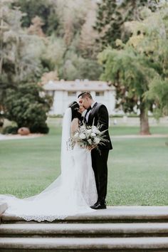 a bride and groom kissing on the steps in front of their wedding venue, with trees behind them