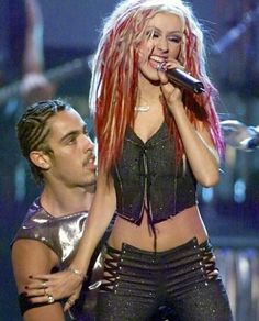 lady with red hair singing on stage next to a man in black pants and crop top