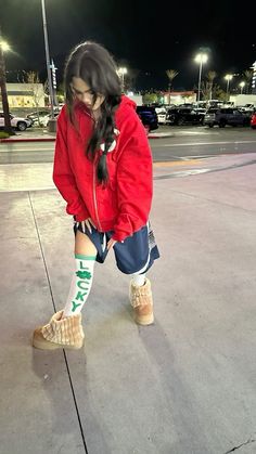 a girl in red jacket and blue shorts standing on street next to parking lot at night