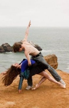 two people standing on top of a cliff next to the ocean with their arms in the air