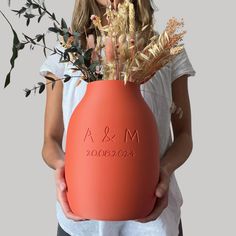 a woman holding an orange vase with flowers in it and the words a & m written on it