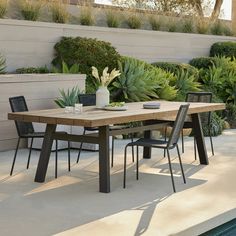 a wooden table sitting next to a pool with chairs around it and plants in the background