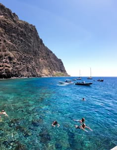 people swimming in clear blue water next to a rocky cliff face with boats on it