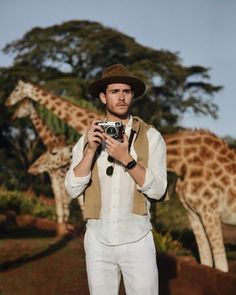 a man in white pants and a hat holding a camera near two giraffes