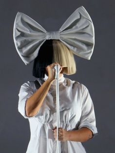 a woman wearing a white shirt and bow tie holding a microphone to her face while standing in front of a gray background