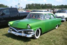 an old green car is parked in the grass near other antique cars and pickup trucks