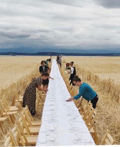 several people standing around a long table in the middle of a wheat field with glasses on it