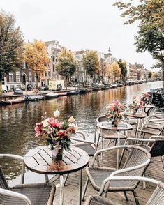 tables and chairs on the side of a river with boats in the water behind them