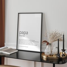 a black table topped with a framed poster next to a vase filled with flowers and an open book