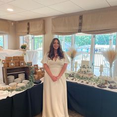 a woman in a white dress standing next to a table with cakes and desserts