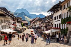 many people are walking down the cobblestone street in an old european town with mountains in the background