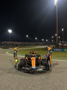 two men standing next to a racing car on a race track at night with lights in the background