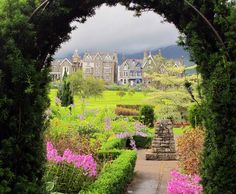 an archway leading into a lush green garden with pink flowers in the foreground and houses in the background