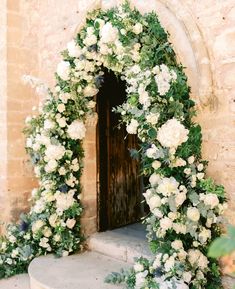 an arch covered in white flowers and greenery next to a doorway with a wooden door