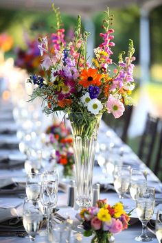 a vase filled with colorful flowers sitting on top of a long table covered in glasses