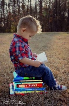 a young boy sitting on top of a stack of books in the grass with trees behind him