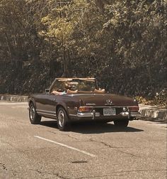 a brown convertible car driving down a street next to a lush green tree filled forest