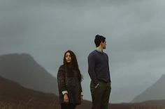 two people standing in tall grass with mountains in the background and cloudy sky