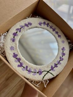 a round mirror with purple flowers on it in a cardboard box next to a window