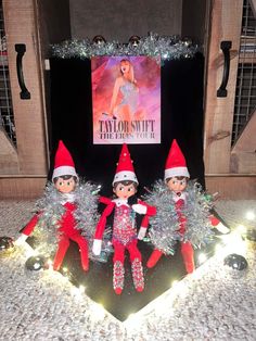 three elfs sitting in front of a sign with christmas lights on the bottom and an advertisement behind them