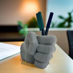 a hand holding two pens on top of a desk next to an open book and pen holder