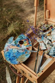 an artist's easel with paint and brushes in it on the ground next to flowers