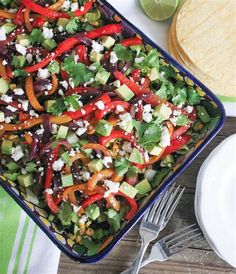 a colorful salad is in a blue dish with tortillas and limes on the side