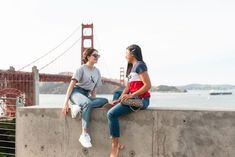 two young women sitting on the edge of a cement wall looking at each other in front of the golden gate bridge