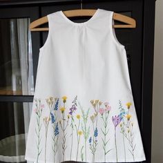 a white dress with flowers painted on the front and back, hanging from a wooden hanger