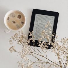 a cup of coffee next to an open book on a table with flowers and a tablet