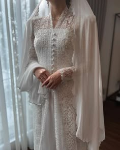 a woman in a white dress and veil standing next to a window with sheer curtains