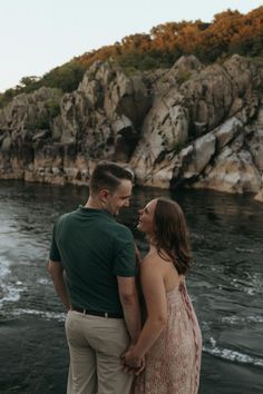 a man and woman standing next to each other by the water looking at each other