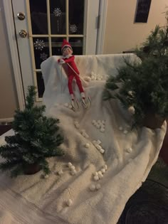 an elf is sitting on top of a blanket with snow around it and trees in front of the door