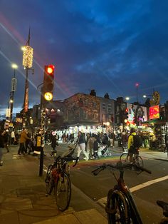 many people are walking and riding bikes on the street at night time with lights in the background