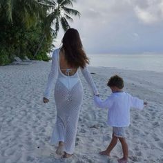 a woman walking on top of a sandy beach next to a little boy holding hands