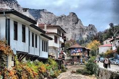 people are walking down the street in front of some buildings and mountains with trees on both sides