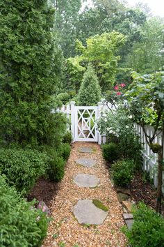 a white gate surrounded by green trees and graveled path with stepping stones leading to it