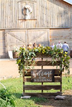 a wooden sign that says welcome to the wedding with greenery on it and people in the background