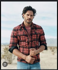 a man standing in the desert with his hand on his hip wearing a red and black plaid shirt