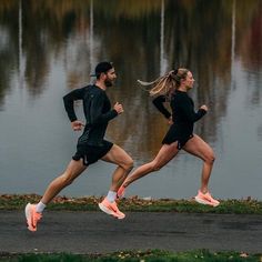 two people are running in the street near some water with trees and grass behind them