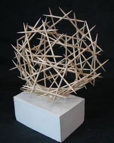 a sculpture made out of sticks sitting on top of a white square block in front of a black background