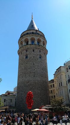 people walking around in front of a tall tower with a red object on it's side