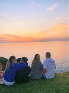 four people are sitting on the grass by the water watching the sun set over the ocean