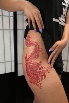 a woman is showing off her red dragon tattoo on her thigh while standing in front of a tiled wall