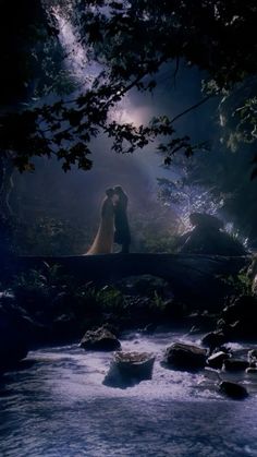 a man and woman are standing on a bridge over a stream in the woods at night