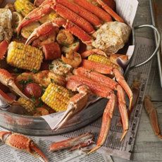 crab legs, corn on the cob and potatoes in a bowl with newspaper next to it