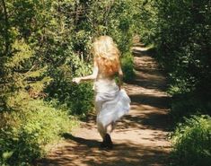 a woman in white dress walking down a dirt road surrounded by green bushes and trees