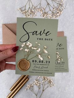 a person holding up a save the date card next to some gold and white flowers