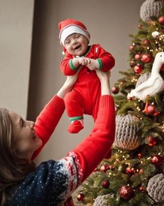 a woman holding up a baby in front of a christmas tree with decorations on it